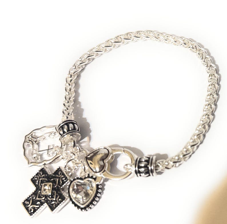 May include: Silver-toned charm bracelet featuring a woven chain design. The bracelet includes various charms: a cross with a central gem, a heart-shaped charm with a clear stone, and a heart-shaped charm. The bracelet has a clasp closure.