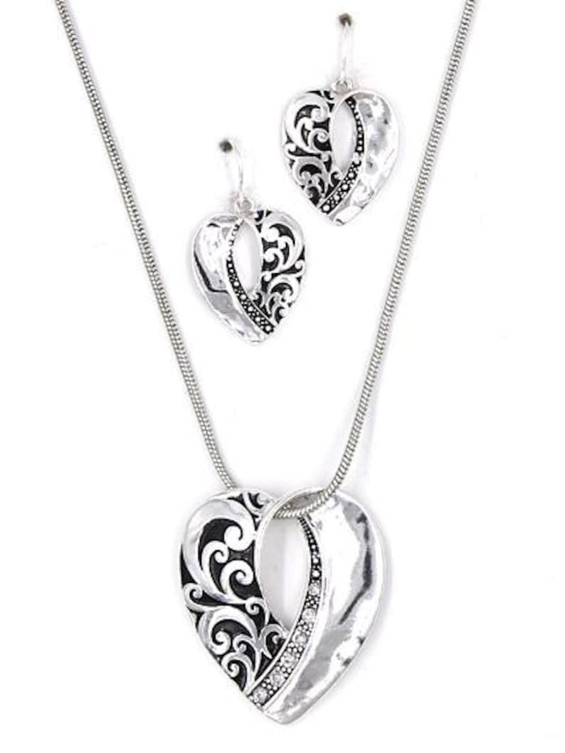 K&ouml;nnte beinhalten: Ein silberfarbenes Schmuckset mit einer herzf&ouml;rmigen Anh&auml;ngerkette und passenden Ohrringen. Der Anh&auml;nger und die Ohrringe haben eine geh&auml;mmerten Textur mit einem schwarzen Filigranmuster und kleinen klaren Steinen.