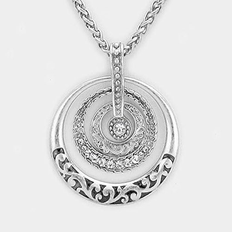 Puede incluir: Collar de plata con un colgante circular que presenta tres c&iacute;rculos conc&eacute;ntricos con un cristal en el centro. El c&iacute;rculo exterior tiene un dise&ntilde;o floral decorativo.