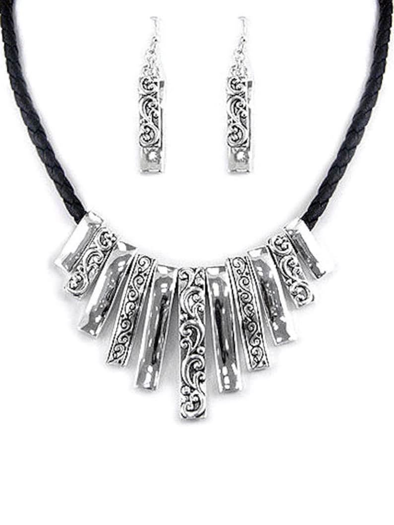 Może przedstawiać: Srebrny zestaw biżuterii składający się z naszyjnika i pasujących kolczyk&oacute;w. Naszyjnik ma czarny pleciony sznurek i wisiorek w kształcie wachlarza z ozdobnymi zawijasami. Kolczyki są prostokątne z podobnymi detalami.