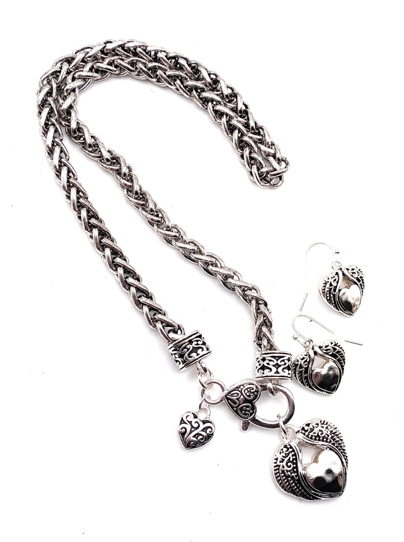 May include: Silver necklace with a braided chain and a heart pendant with wings. The necklace comes with matching earrings with heart pendants and wings.