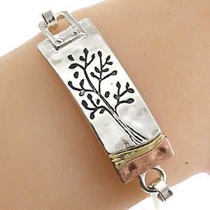 May include: Silver bracelet with a rectangular charm featuring a tree design. The charm has a gold and copper accent.