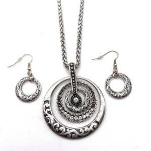 Puede incluir: Un collar de plata con un colgante que presenta tres c&iacute;rculos conc&eacute;ntricos con un dise&ntilde;o decorativo y brillantes de cristal transparentes. Pendientes de plata a juego con un colgante de c&iacute;rculo &uacute;nico.