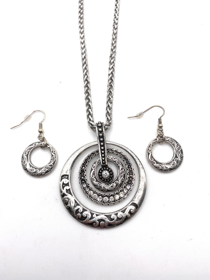 Puede incluir: Un collar de plata con un colgante que presenta tres c&iacute;rculos conc&eacute;ntricos con un dise&ntilde;o decorativo y brillantes de cristal transparentes. Pendientes de plata a juego con un colgante de c&iacute;rculo &uacute;nico.