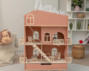 May include: A pink wooden dollhouse with multiple levels, windows, and a staircase. The dollhouse has a drawer at the bottom and is supported by wooden legs. The word "Dollhouse" is written on the roof. The dollhouse is decorated with miniature furniture.