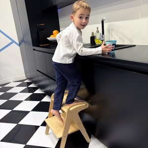 Peut inclure: Un jeune enfant debout sur un marchepied en bois dans une cuisine. L'enfant porte une chemise blanche et un pantalon bleu. L'enfant tend la main vers un comptoir. Le comptoir est noir et a un évier blanc. Il y a des fruits oranges dans un bol sur le comptoir.