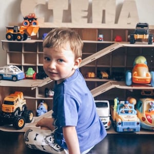 Puede incluir: Un ni&ntilde;o peque&ntilde;o se sienta frente a una estanter&iacute;a de juguetes de madera con el nombre "STELLA" escrito en letras grandes de madera. La estanter&iacute;a est&aacute; llena de coches y camiones de juguete, incluyendo un coche de polic&iacute;a, un cami&oacute;n volquete amarillo y una camioneta azul.