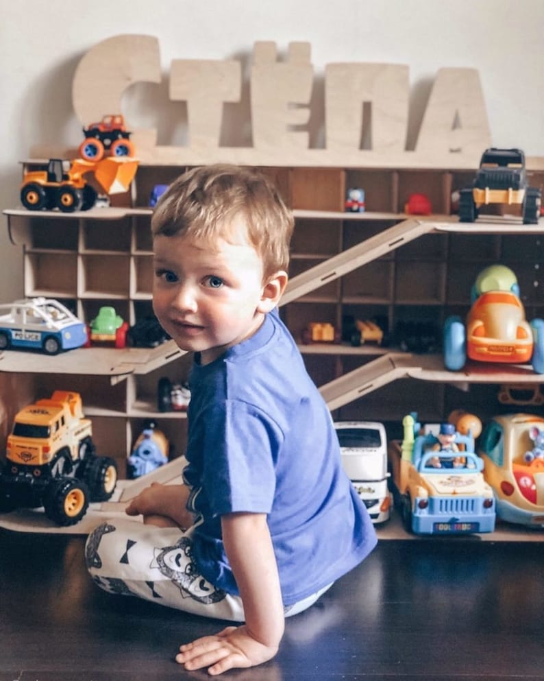 Puede incluir: Un ni&ntilde;o peque&ntilde;o se sienta frente a una estanter&iacute;a de juguetes de madera con el nombre "STELLA" escrito en letras grandes de madera. La estanter&iacute;a est&aacute; llena de coches y camiones de juguete, incluyendo un coche de polic&iacute;a, un cami&oacute;n volquete amarillo y una camioneta azul.