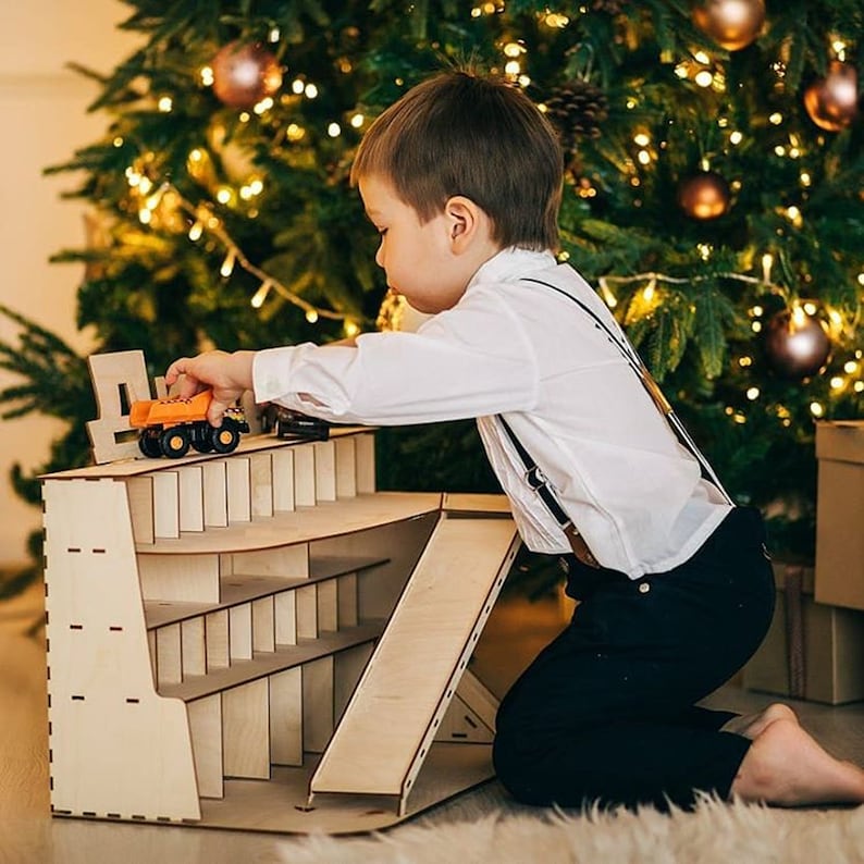 May include: A child plays with a toy truck and a wooden toy garage with a ramp. The garage has multiple shelves for storing toys. The child is kneeling on a white rug in front of a Christmas tree.