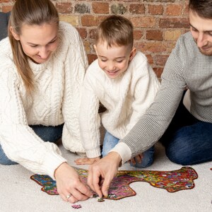 Op de afbeelding: Een gezin van drie werkt samen om een ​​kleurrijke puzzel op een wit tapijt te voltooien. De puzzelstukjes zijn verspreid rond de geassembleerde puzzel. Het gezin draagt ​​casual kleding en glimlacht.