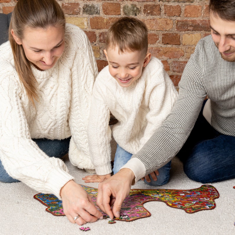 Op de afbeelding: Een gezin van drie werkt samen om een ​​kleurrijke puzzel op een wit tapijt te voltooien. De puzzelstukjes zijn verspreid rond de geassembleerde puzzel. Het gezin draagt ​​casual kleding en glimlacht.