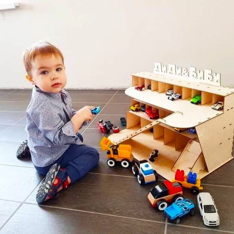 May include: A wooden toy car garage with multiple levels and a ramp. The garage has a sign that says "Диди & Биби". A young child is playing with toy cars in front of the garage.