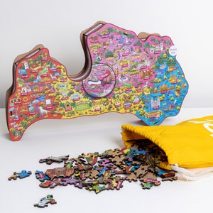 Op de afbeelding: Een kleurrijke houten puzzel van Letland, met een kaart van het land en verschillende bezienswaardigheden. De puzzel is gedeeltelijk in elkaar gezet, met wat stukjes die rond een gele trekkoordtas zijn verspreid. De tas heeft het woord "Atvērt" erop gedrukt.