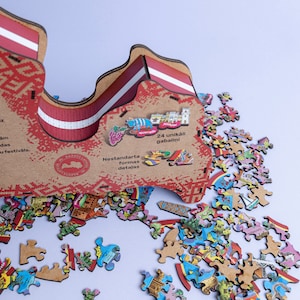 Op de afbeelding: Een houten puzzel van Letland, met 24 unieke stukjes. De puzzel heeft de vorm van Letland en heeft een rood-wit gestreept ontwerp. De puzzel is gedeeltelijk in elkaar gezet, met enkele stukjes die ontbreken. De tekst "24 unikāli gabalīni" is zichtbaar op de puzzeldoos.