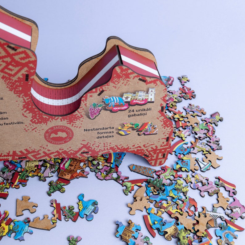 Op de afbeelding: Een houten puzzel van Letland, met 24 unieke stukjes. De puzzel heeft de vorm van Letland en heeft een rood-wit gestreept ontwerp. De puzzel is gedeeltelijk in elkaar gezet, met enkele stukjes die ontbreken. De tekst "24 unikāli gabalīni" is zichtbaar op de puzzeldoos.
