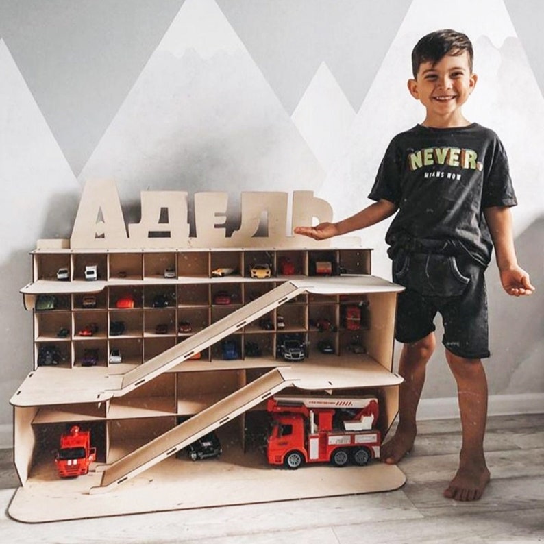 Puede incluir: Un garaje de madera para coches de juguete con una rampa y varios niveles. El garaje est&aacute; etiquetado con el nombre "ADELL" en letras grandes de madera. Un ni&ntilde;o peque&ntilde;o est&aacute; de pie junto al garaje.