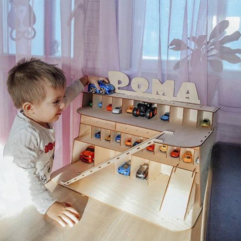 May include: A wooden toy car garage with a ramp and multiple levels for storing toy cars. The garage is personalized with the name "POMA" and features a blue toy car on the top level.