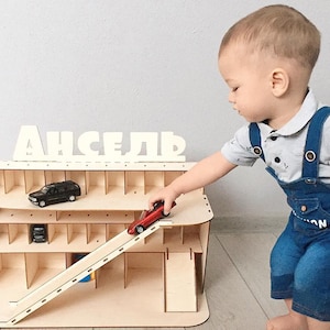 May include: A wooden toy garage with a ramp and multiple levels for storing toy cars. The garage has a sign that says "АНСЕЛЬ" in white letters. A young child is playing with a red toy car on the ramp.