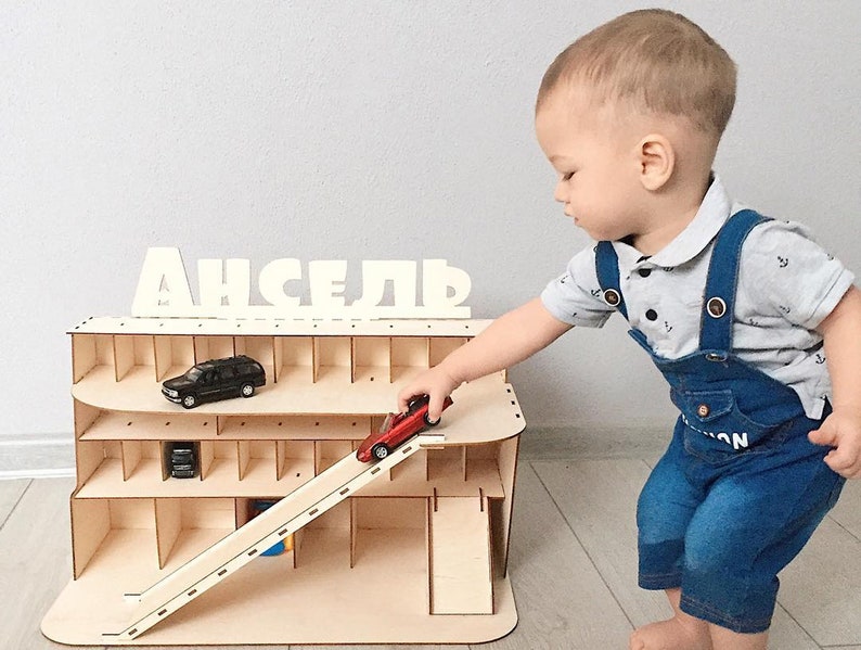 May include: A wooden toy garage with a ramp and multiple levels for storing toy cars. The garage has a sign that says "АНСЕЛЬ" in white letters. A young child is playing with a red toy car on the ramp.