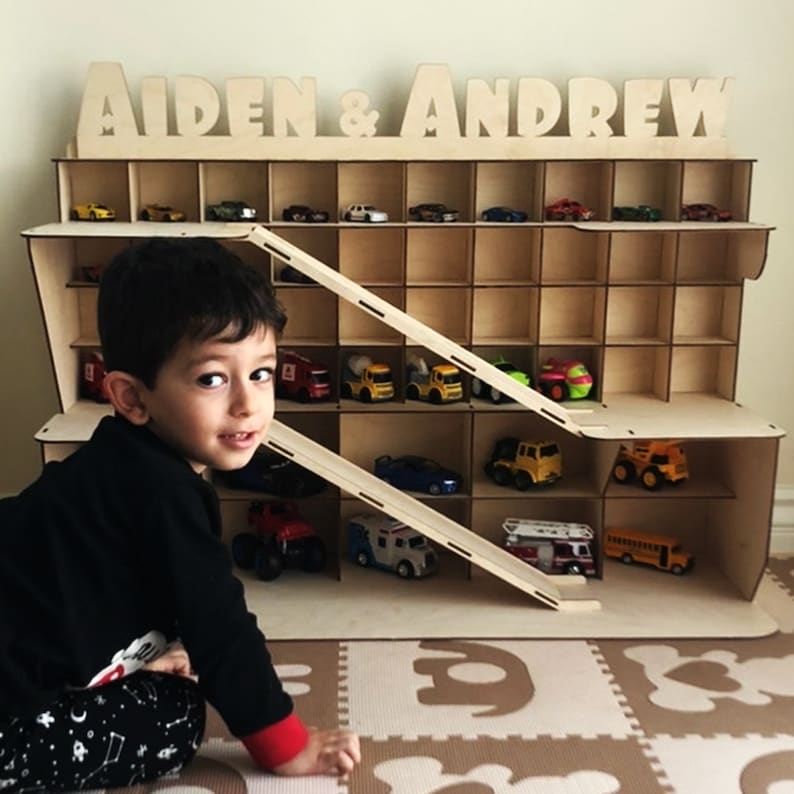 Puede incluir: Un garaje de madera para coches de juguete con una rampa y estantes. El garaje est&aacute; etiquetado como "Aiden & Andrew". Un ni&ntilde;o peque&ntilde;o est&aacute; sentado frente al garaje.