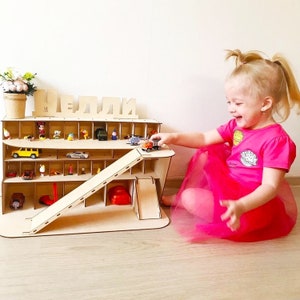 May include: A wooden toy car garage with a ramp and multiple shelves for storing toy cars. The garage is labeled "HELLI" and has a variety of toy cars and figures on the shelves. A young child is playing with the garage and cars.