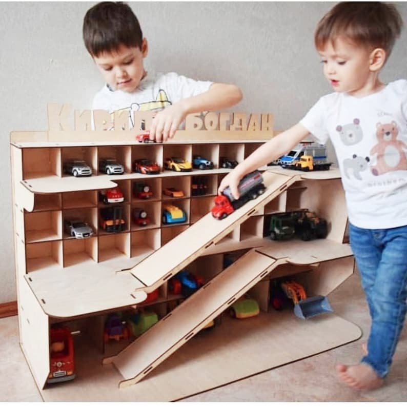 Puede incluir: Un garaje de juguete de madera con m&uacute;ltiples estantes y dos rampas para coches de juguete. El garaje est&aacute; etiquetado con "Кирилл Богдан" en letras cir&iacute;licas. Dos ni&ntilde;os est&aacute;n jugando con coches de juguete en el garaje.