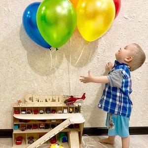 May include: A wooden toy car garage with a ramp and a helicopter on top. The garage has multiple levels and spaces for toy cars. A young child is reaching up towards a cluster of colorful balloons.
