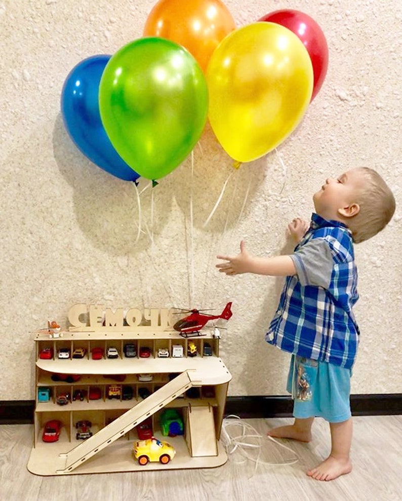 May include: A wooden toy car garage with a ramp and a helicopter on top. The garage has multiple levels and spaces for toy cars. A young child is reaching up towards a cluster of colorful balloons.