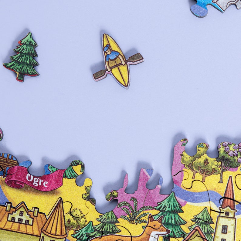 Op de afbeelding: Een kleurrijk puzzelstuk met een gele kano en een persoon die peddelt, een groene boom en een deel van een grotere puzzel met een stadssc&egrave;ne, waaronder een gebouw met het woord "Ogre" op een banner.