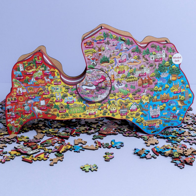 Op de afbeelding: Een kleurrijke puzzel van Letland, met een kaart van het land en verschillende bezienswaardigheden en illustraties. De puzzel is gedeeltelijk in elkaar gezet, met wat stukjes die rond de hoofdafbeelding zijn verspreid. De tekst "Atvērt Seit" is zichtbaar op de puzzel.