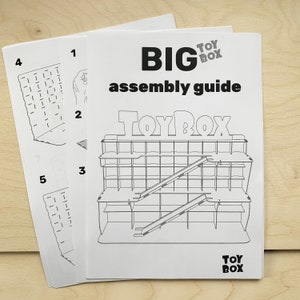 Puede incluir: Gu&iacute;a de montaje para una gran caja de juguetes de madera con una rampa. La gu&iacute;a muestra los pasos para construir la caja de juguetes, que tiene un gran letrero de "Toy Box" en la parte delantera. La gu&iacute;a se titula "BIG TOY BOX assembly guide".