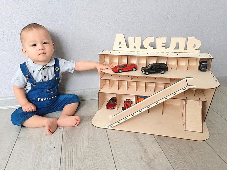 May include: A wooden toy car garage with a ramp and multiple levels. The garage is labeled "ANSElR" and has several toy cars inside. A young child is sitting on the floor in front of the garage.