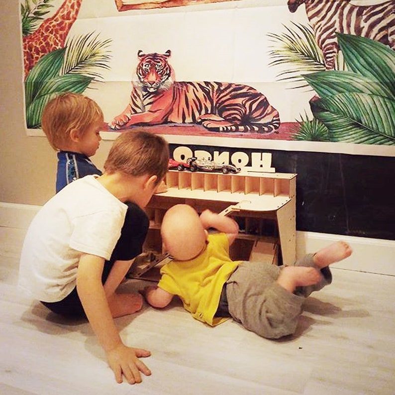 May include: Three children play with a wooden toy car garage. The garage has multiple levels and slots for toy cars. The children are sitting on a light wood floor. A poster with a tiger and zebra is on the wall behind them.