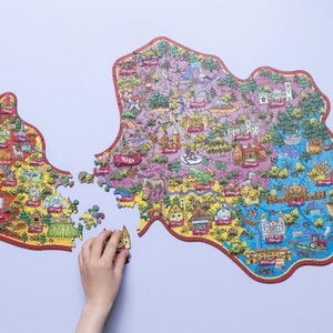 Op de afbeelding: Een kleurrijke puzzel die een kaart van Riga, Letland, voorstelt. De puzzelstukken zijn gevormd naar de omtrek van de stad en bevatten verschillende bezienswaardigheden en illustraties. De puzzel is gedeeltelijk in elkaar gezet, sommige stukken ontbreken.