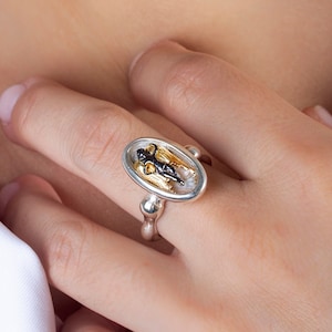 May include: Oval-shaped silver ring with a detailed black and gold design. The ring is worn on a finger, showcasing the intricate craftsmanship. The ring's design is set against a silver band.