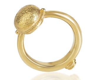 May include: A gold ring featuring a textured, oval-shaped centrepiece. The ring has a smooth, rounded band and a small, decorative detail on the side. The ring is made of a shiny, yellow metal.