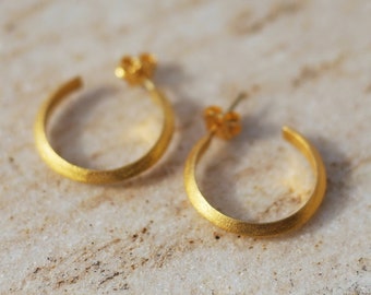 May include: A pair of gold-colored hoop earrings. The earrings have a textured, matte finish and a simple, circular design. The earrings are displayed on a light-colored, marble-like surface. The earrings are small and delicate, suitable for everyday wear.