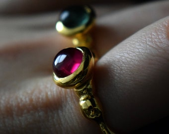May include: Close-up of two gold rings, each featuring a gemstone. The lower ring has a vibrant red gemstone, while the upper ring showcases a green gemstone. The rings have a unique, organic design.