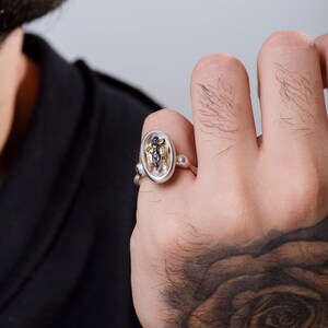 May include: Silver ring featuring an oval-shaped centerpiece with a detailed figure. The ring has a unique design with a combination of silver and gold tones, showcasing intricate craftsmanship. The ring is worn on a hand.