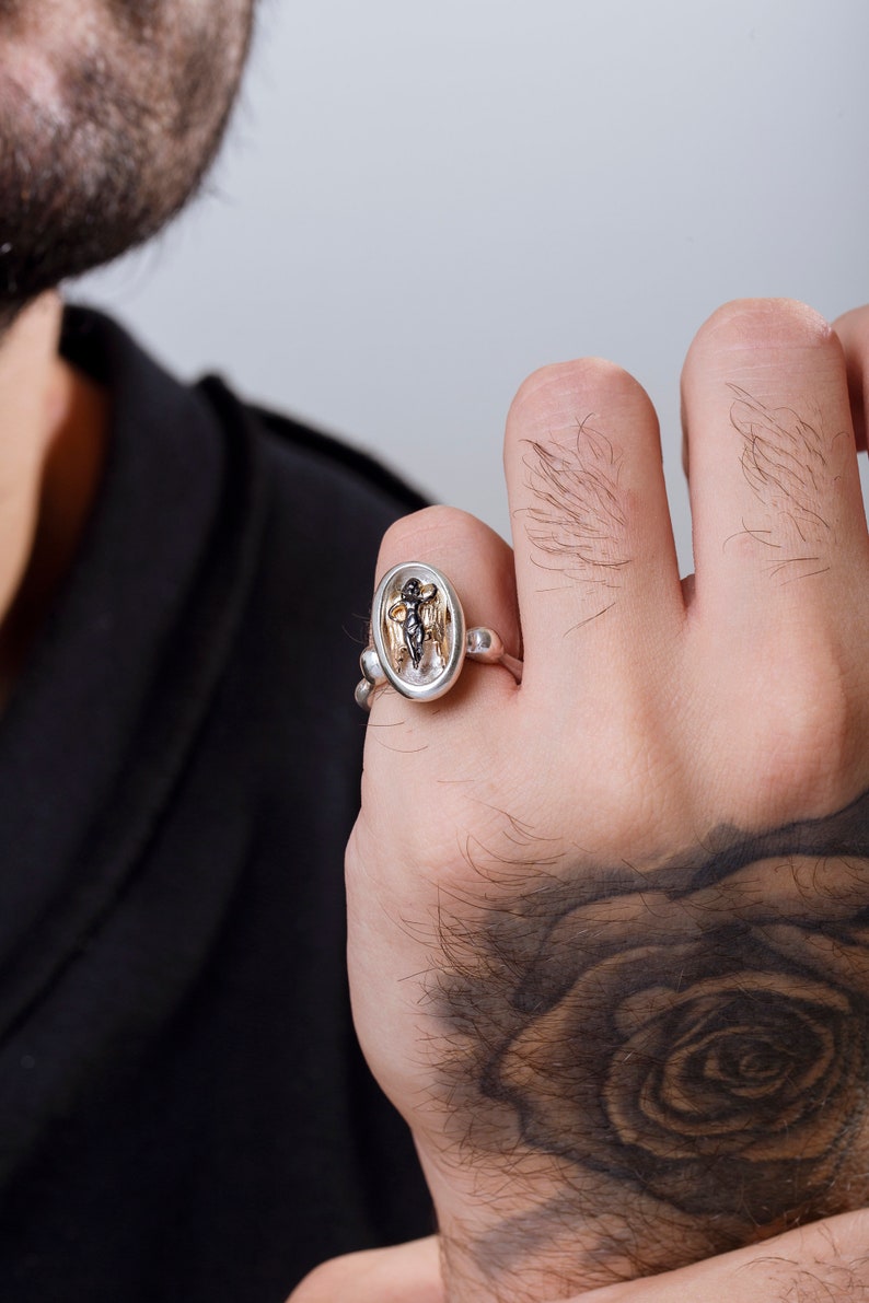 May include: Silver ring featuring an oval-shaped centerpiece with a detailed figure. The ring has a unique design with a combination of silver and gold tones, showcasing intricate craftsmanship. The ring is worn on a hand.