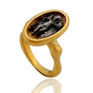 May include: A gold-toned ring featuring an oval-shaped centerpiece with a dark, detailed relief of a cherub. The ring has a smooth, polished finish and a unique, sculpted band. A piece of jewelry with an antique or classical aesthetic.