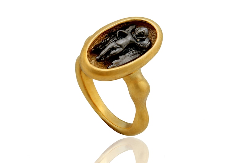 May include: A gold-toned ring featuring an oval-shaped centerpiece with a dark, detailed relief of a cherub. The ring has a smooth, polished finish and a unique, sculpted band. A piece of jewelry with an antique or classical aesthetic.