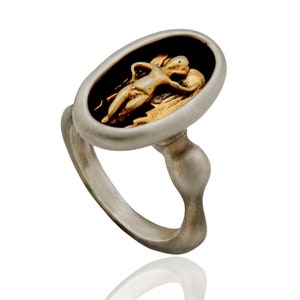 May include: A silver ring with an oval face featuring a gold-colored relief of a reclining figure against a black background. The ring has a unique, curved design.