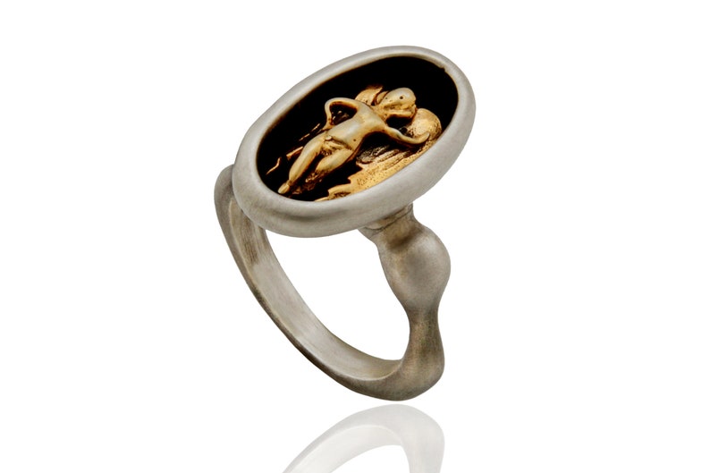 May include: A silver ring with an oval face featuring a gold-colored relief of a reclining figure against a black background. The ring has a unique, curved design.
