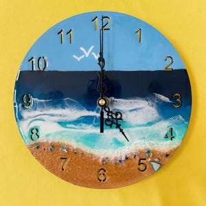 May include: Round wall clock with a beach scene design. The clock face features a blue sky, dark blue ocean, white waves, and a sandy beach. Gold-colored numerals mark the hours. The clock hands are black.