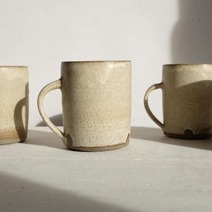 May include: Four beige ceramic mugs and a tumbler are arranged on a white surface. The mugs have handles and a textured appearance, with a darker brown rim and base. The tumbler lacks a handle. The lighting creates shadows.