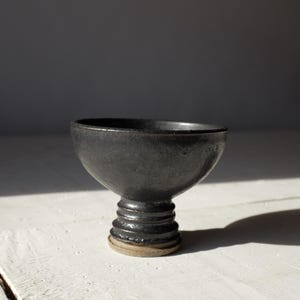 May include: A handmade, black ceramic chalice with a textured surface. The chalice features a rounded bowl shape atop a stem with a series of horizontal ridges. The base is a lighter, unglazed clay color. The chalice is displayed on a white surface.