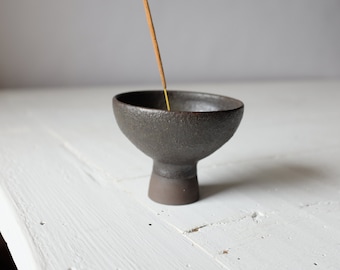 May include: A dark brown ceramic incense holder with a wide bowl and a short, sturdy base. An incense stick is placed in the bowl.