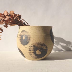 Puede incluir: Un jarrón de cerámica beige con patrones circulares de color gris oscuro. El jarrón está lleno de follaje marrón seco. El jarrón tiene una textura moteada y una forma redondeada. El fondo es una superficie blanca lisa.