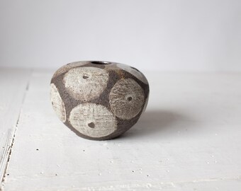 Peut inclure: Un petit vase rond en céramique brune avec un motif géométrique blanc. Le vase a une surface légèrement irrégulière et une large ouverture en haut.